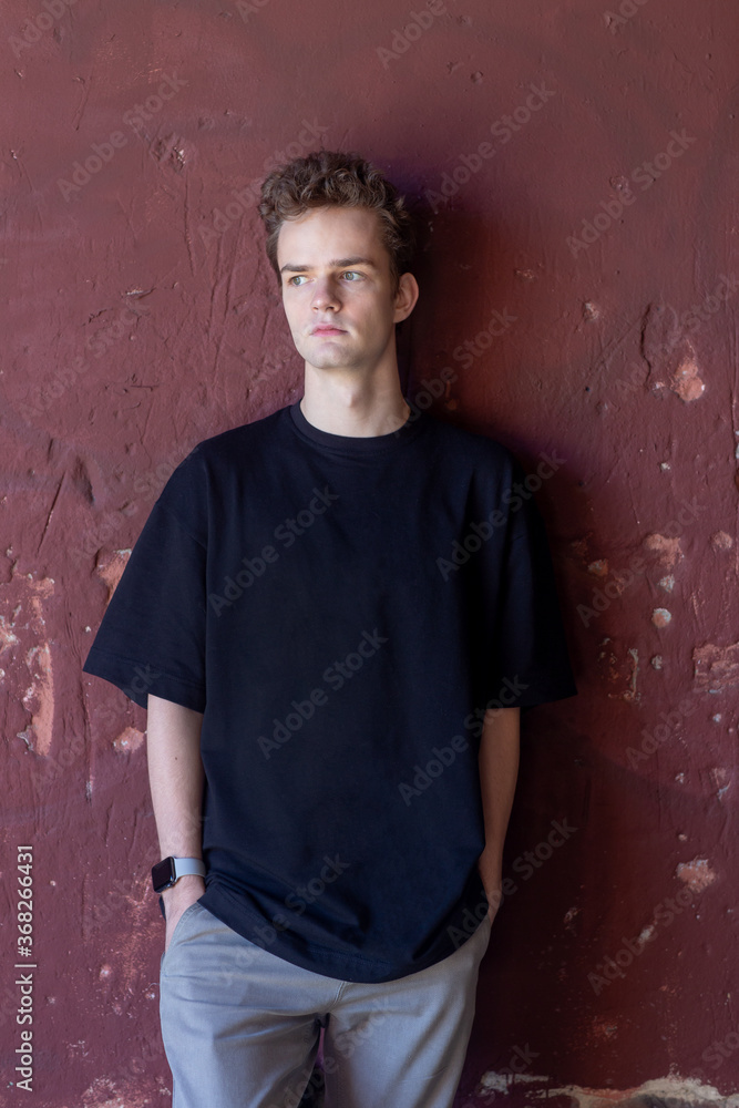 a young guy, a teenager in a dark, black T-shirt stands against a wall background. t-shirt advertising concept a young guy, a teenager in a dark, black T-shirt stands against a wall background. t-shirt advertising concept