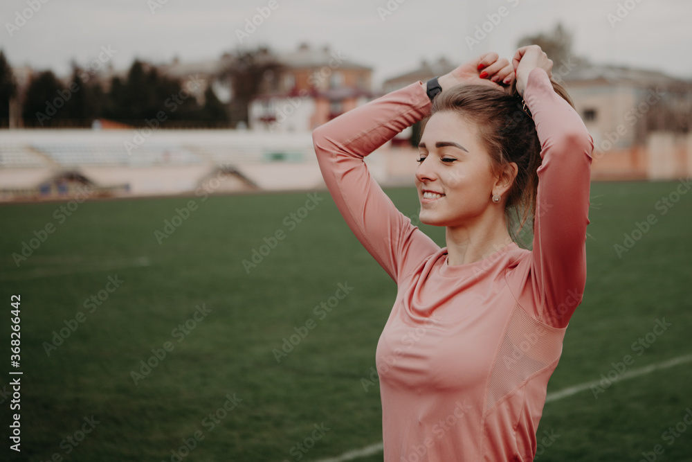 Close up portrait of a girl straightening her hair and smiling. Sporty woman enjoying life and lead healthy lifestyle. Lady training at the city stadium. Image with copy space.