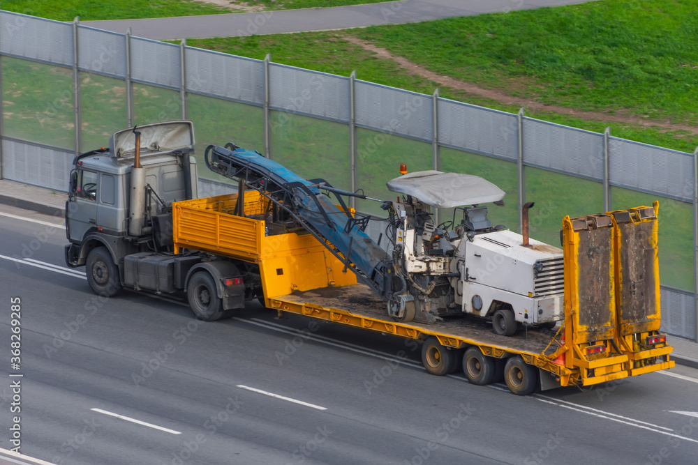 Obraz premium Transportation of equipment for cutting and removing old asphalt pavement for road repair on a truck platform of a truck trailer on a highway in a city, side aerial view.