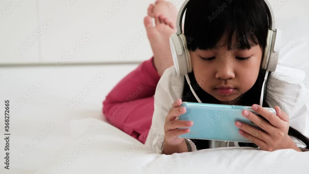 Asian child girl are addictive playing mobile phones with wearing headphones in bedroom Asian child girl are addictive playing mobile phones with wearing headphones in bedroom