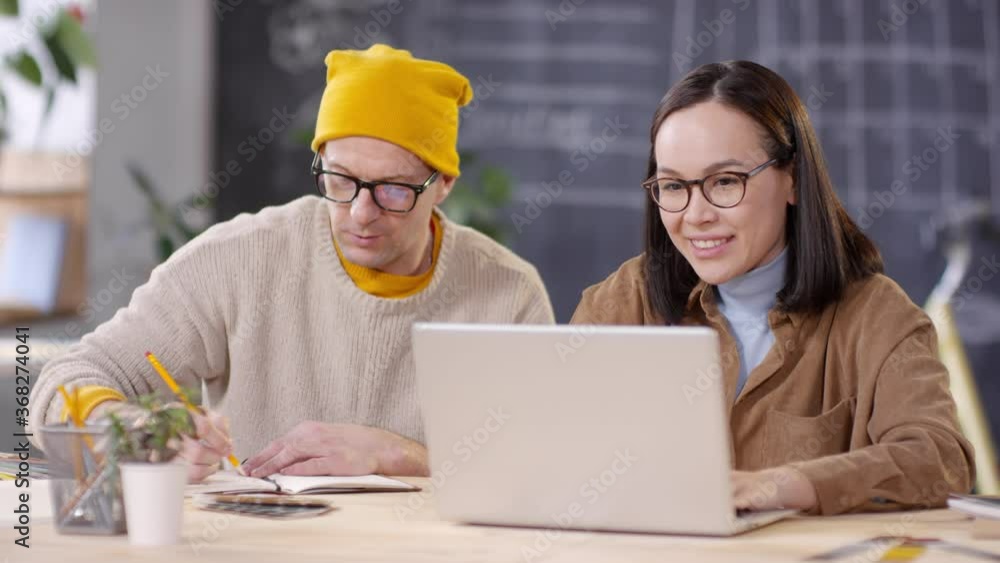 Tracking of middle-aged male designer in glasses and yellow beanie hat sitting at desk with his female colleague and discussing project. He is pointing at something on laptop screen and making notes Tracking of middle-aged male designer in glasses and yellow beanie hat sitting at desk with his female colleague and discussing project. He is pointing at something on laptop screen and making notes