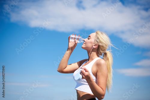Young blond fit woman, wearing black leggings and white top,drinking water from glass bottle outside with blue sunny sky on background. Female fitness training in summer. Healthy lifestyle concept.