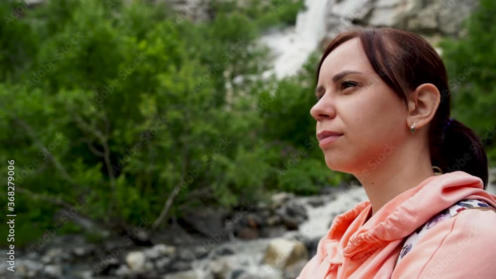 Portrait of young woman looking away on background of waterfall. Adult female tourist enjoying beautiful view in mountainous area. Portrait of young woman looking away on background of waterfall. Adult female tourist enjoying beautiful view in mountainous area.