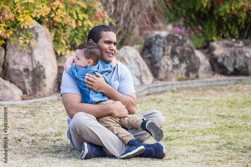 Aboriginal father sitting on grass hugging mixed race aboriginal caucasian son Stock Photo