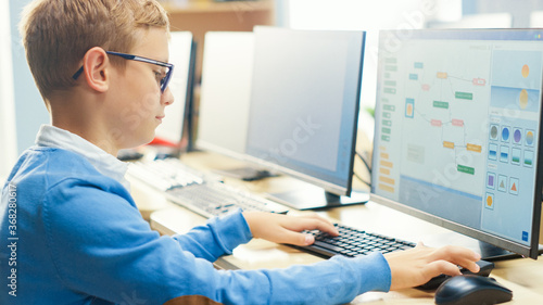 Elementary School Computer Science Classroom: Cute Little Boy wearing Glasses Uses Personal Computer, Learning Programming Language for Software Coding, Over the Shoulder Shot © Gorodenkoff