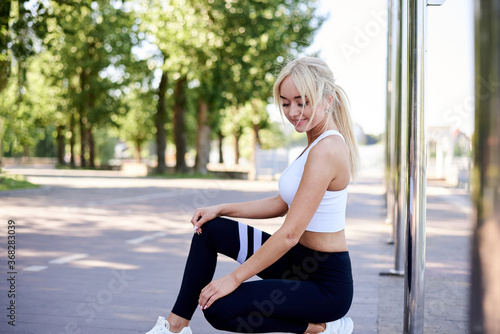 Young fit blond woman, wearing white top and black leggings, squatting down, posing by city lake in park in summer. Sportswoman, resting relaxing after fitness exercise training. Healthy lifestyle. © Natalia