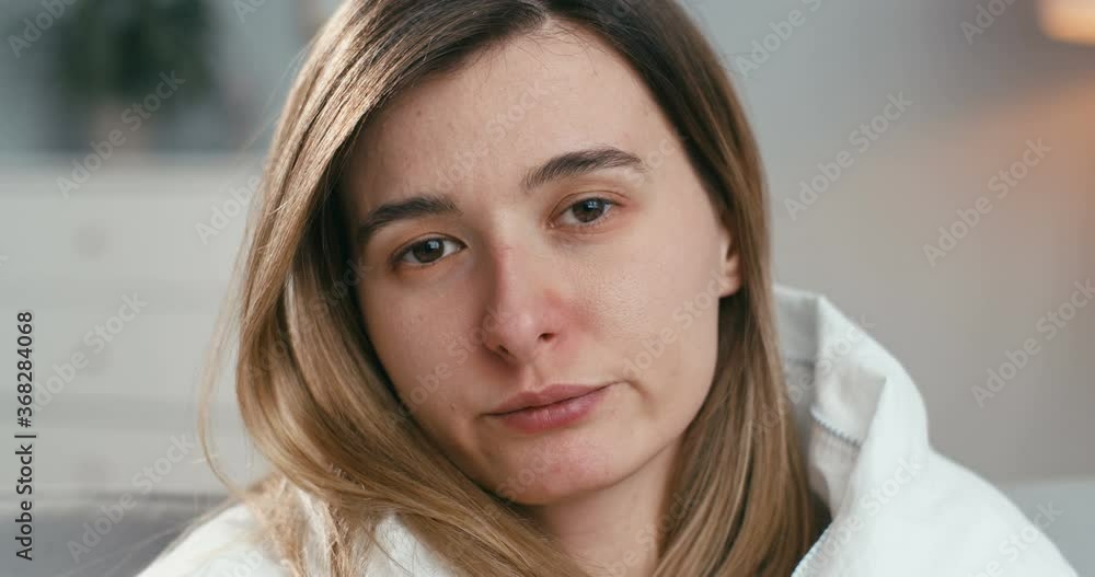 Close up of ill young Caucasian woman looking at camera and blowing nose in napkin while sitting at isolation at home. Portrait of unhealthy girl with flu or coronavirus. Close up of ill young Caucasian woman looking at camera and blowing nose in napkin while sitting at isolation at home. Portrait of unhealthy girl with flu or coronavirus.