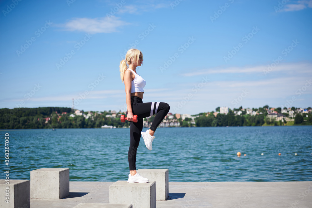 Young blond fit woman, wearing black leggings and white top, training outside by city lake on concrete platform, holding red weights. Female power training in summer. Healthy lifestyle concept.