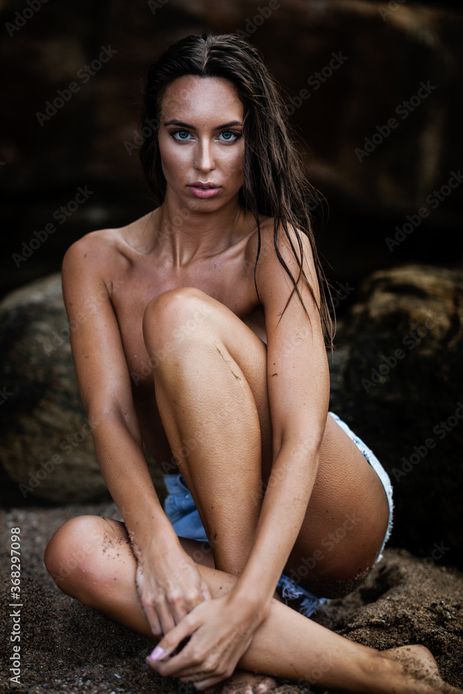 Summertime recreation concept. Beautiful young sexy woman with fit trained slim body wearing blue shorts sits on a beach on a stone. Fashion female model poses by the sea