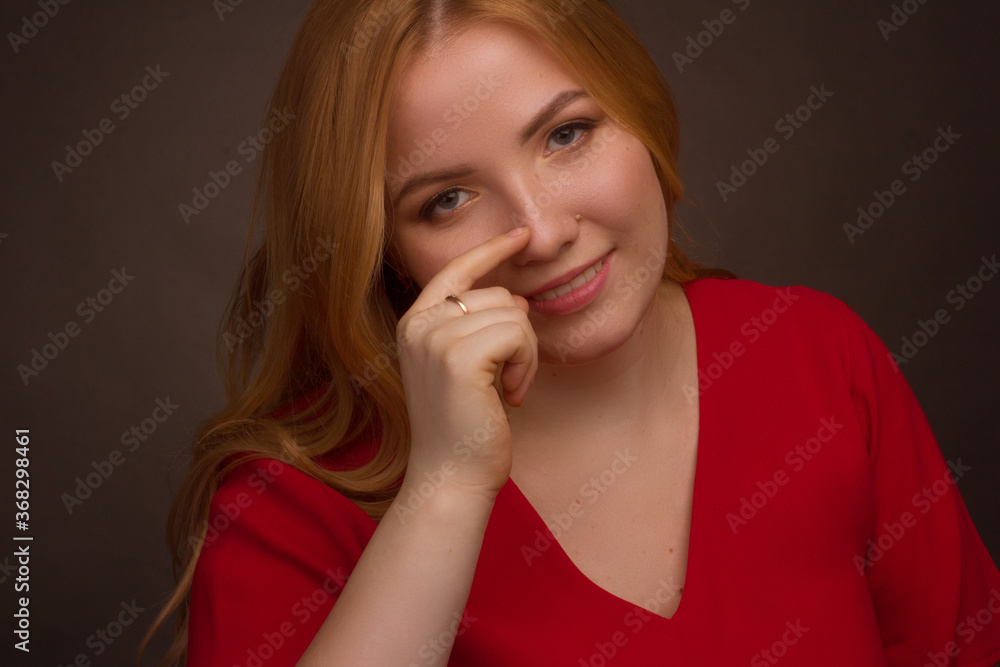 Studio portrait of an overweight young woman with light hair in a red dress on a black background. he scratches his nose with his finger Studio portrait of an overweight young woman with light hair in a red dress on a black background. he scratches his nose with his finger