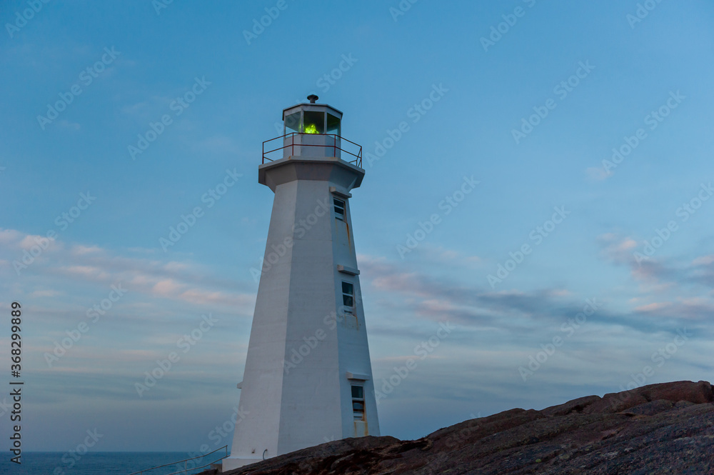 Lighthouse tower made of white concrete hexagonal, six-sided tapering concrete. The structure stands on a rock cliff. The lighthouse has a blue sky with clouds. There's a green lantern in the cupola. 