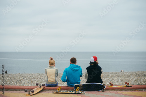 Friends are relax on the embankment of the Black Sea after riding on longboards. They chat and laugh fun with the view of the sea and pebble beach with fishermen