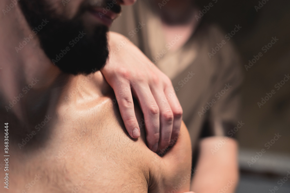Fototapeta premium close-up male masseur does a sports shoulder massage to a muscular male athlete in a room with a contrasting dark light. Professional sports massage
