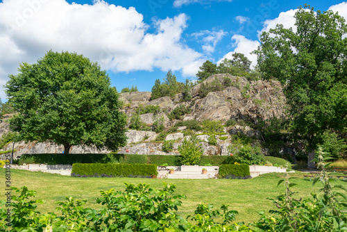 Fototapeta Naklejka Na Ścianę i Meble -  Landscape design in Scandinavia at sunny summer day. Rocky stone cliffs frame the green trees in the garden. Beautiful smooth green mown manicured  lawn. Garden design with trimmed shrubs. 