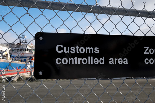 A black and white fence with a sign attached to a chain link industrial boarder. It has customs controlled area in white text. There's a harbour with a wharf in the background under a blue sky.