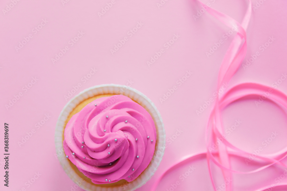 A beautiful cupcake with bright pink cream and a little silver decoration on a pink background. Pink ribbon for decor in the foreground lies in waves. Festive background with free space. A beautiful cupcake with bright pink cream and a little silver decoration on a pink background. Pink ribbon for decor in the foreground lies in waves. Festive background with free space.