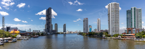 A panoramic view of Bangkok with, the blue sky and white cloud, the Chao Phraya River flowing through, and many tall buildings, skyscraper, and condominiums on both of the riverside.