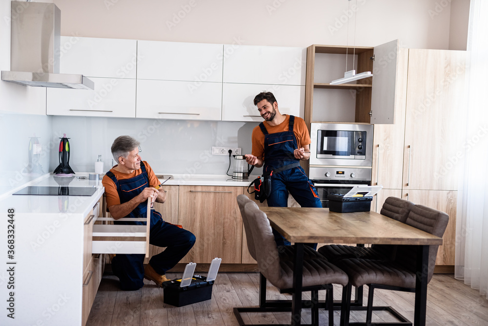Full length shot of two handymen, workers in uniform talking while assembling kitchen cabinet using screwdriver indoors. Furniture repair and assembly concept