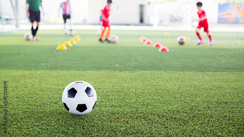 Soccer ball on green artificial turf with blurry kid soccer team training. Blurry kid soccer player jogging between marker cones and control ball with soccer equipment in football academy. © Koonsiri