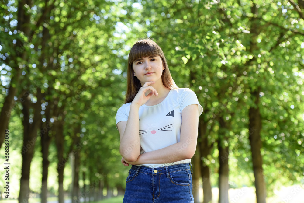 A young girl stands thoughtfully behind a background of green trees . A young girl stands thoughtfully behind a background of green trees .
