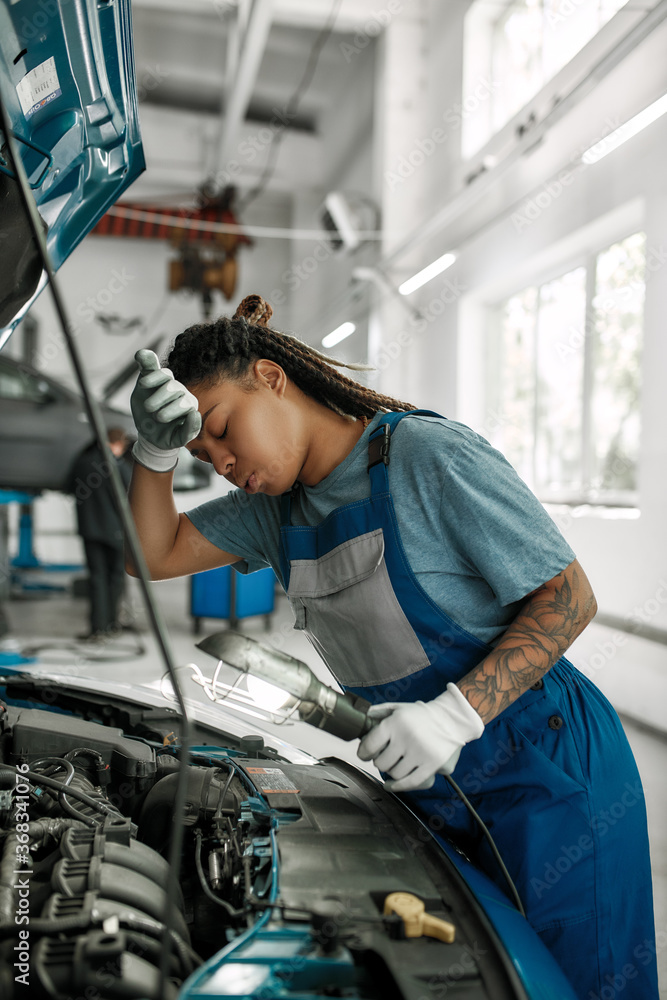 Your car doctor. Young african american woman, professional female mechanic wiping forehead, examining under hood of car with torch at auto repair shop Your car doctor. Young african american woman, professional female mechanic wiping forehead, examining under hood of car with torch at auto repair shop