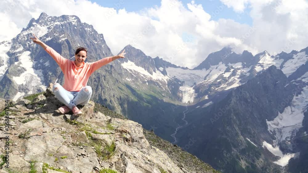 Young happy woman sitting on rock against cloudy sky on sunny day in mountains. Female traveler admires beautiful view in mountainous area. Young happy woman sitting on rock against cloudy sky on sunny day in mountains. Female traveler admires beautiful view in mountainous area.