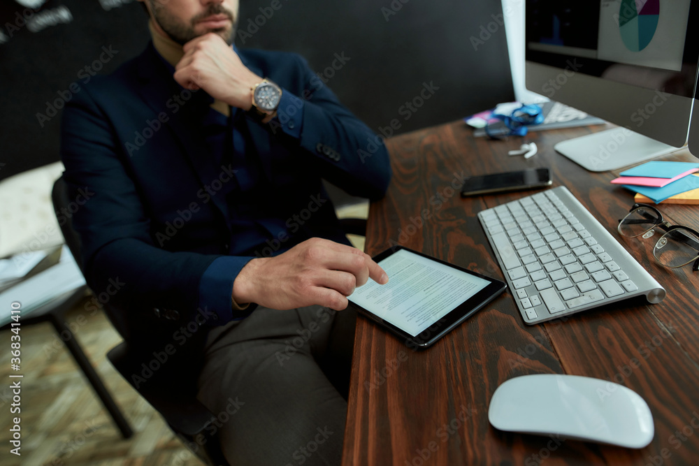 Business and finance concept. Young businessman, sales manager sitting at his workplace in the modern office and reading financial business news online on digital tablet Business and finance concept. Young businessman, sales manager sitting at his workplace in the modern office and reading financial business news online on digital tablet