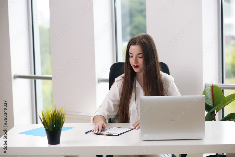 Portrait of young businesswoman wearing white shirt sitting in modern office with laptop Portrait of young businesswoman wearing white shirt sitting in modern office with laptop