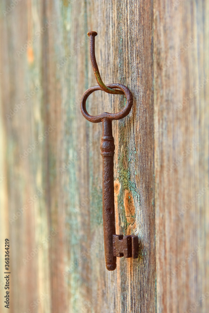 An old forged beautiful rusty key hangs on an old iron nail on a wooden wall, with peeling paint An old forged beautiful rusty key hangs on an old iron nail on a wooden wall, with peeling paint