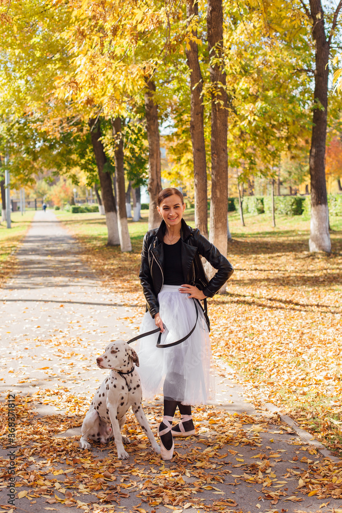Ballerina with Dalmatian dog in the Park. Woman ballerina in a white ballet skirt and black leather jacket dancing in pointe shoes in autumn park with her spotty dalmatian dog. Ballerina with Dalmatian dog in the Park. Woman ballerina in a white ballet skirt and black leather jacket dancing in pointe shoes in autumn park with her spotty dalmatian dog.