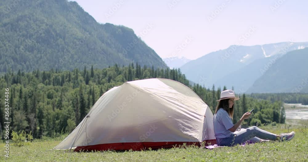 Woman hiker is sitting on mountain near tent and making photos on smartphone admiring scenery view, side view. Traveling, vacation, journey, hiking, camping concept. Blogger is making content. Woman hiker is sitting on mountain near tent and making photos on smartphone admiring scenery view, side view. Traveling, vacation, journey, hiking, camping concept. Blogger is making content.