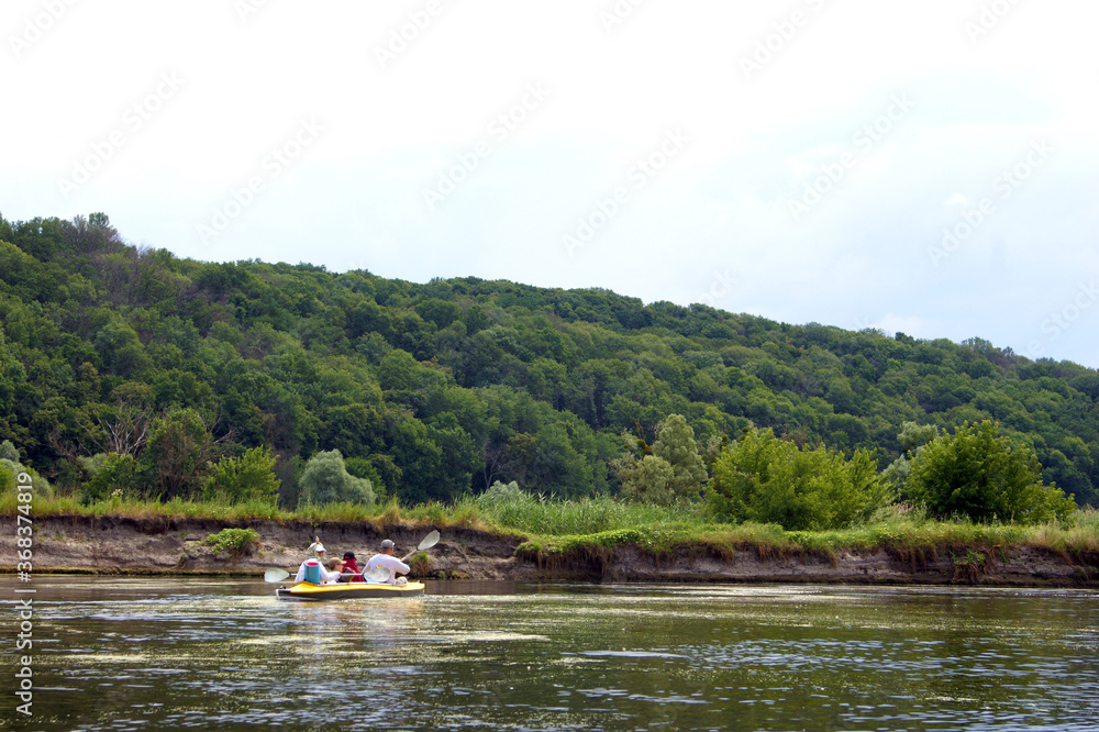 Family rowing in a yellow kayak in summer along the trees at the bank of Seversky Donets river.