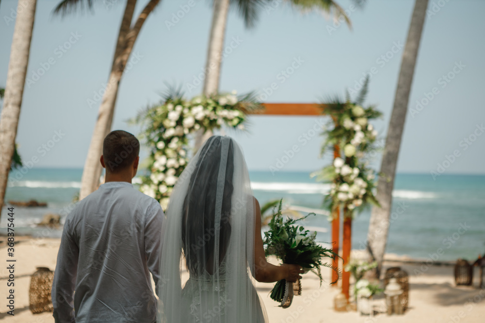 Wedding ceremony on a tropical beach. Happy groom and beautiful blond bride walk to the the arch decorated with flowers. Sea in the background. Wedding ceremony on a tropical beach. Happy groom and beautiful blond bride walk to the the arch decorated with flowers. Sea in the background.