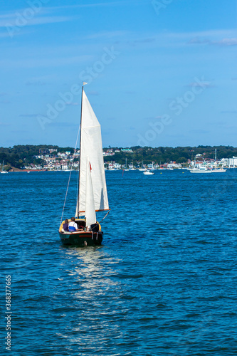 A few people are on a small wooden boat sailing full sails ahead. It is a sunny afternoon and sea is calm. There is a white banner on top of the mast pole.  Reflection is seen in clear waters photo