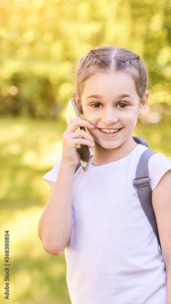 Cute little girl talking. Hold phone near face. Young woman smile. Looking at camera. Outdoor school. Green background. Smiling speach. Happy expression. Student lifestyle. Vertical Cute little girl talking. Hold phone near face. Young woman smile. Looking at camera. Outdoor school. Green background. Smiling speach. Happy expression. Student lifestyle. Vertical