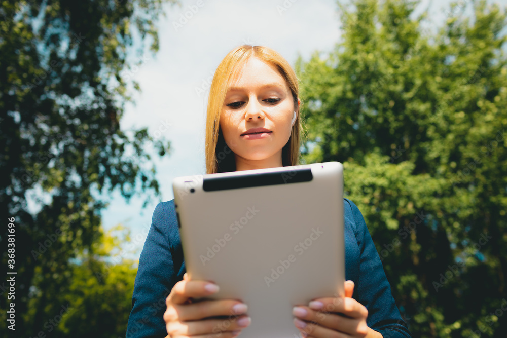 Fototapeta premium Young happy smiling beautiful caucasian adult girl using mobile tablet in the park at summer