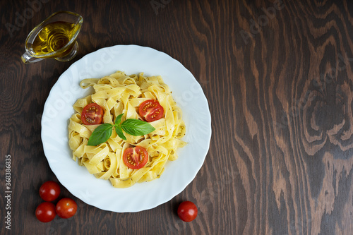 Top view of organic italian fettucine pasta in plate decorated with basil leaves together with olive oil gravy boat and whole tomatoes on dark brown wooden surface. Image with copy space