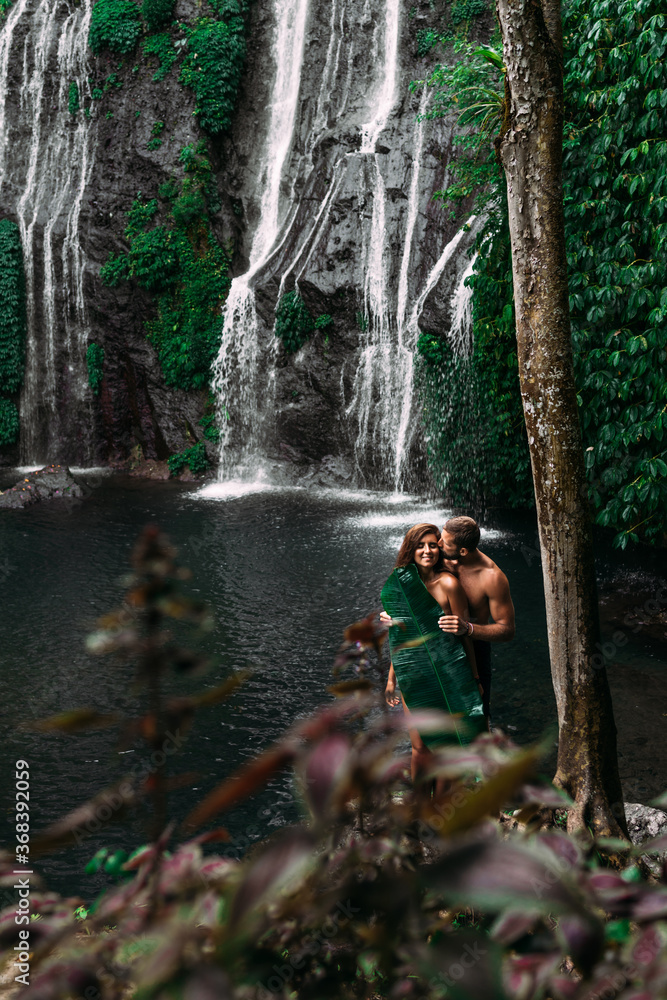 Fototapeta premium Lovers at the waterfall. Beautiful couple at a waterfall in Indonesia. A couple in love travels around the island of Bali. A man and a woman kiss at the waterfall. Travel to Asia. Сopy space
