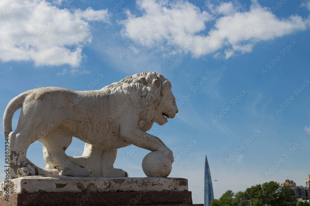 The lion sculpture looks at the Lakhta center, against a blue sky with clouds.