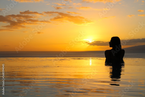 the Girl is standing back in the pool  slightly turning her head  her silhouette is reflected in the water. Against the background of the sunset and the ocean.