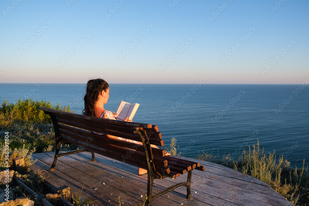 young woman sitting on a bench and reading book outdoors with summer sea background