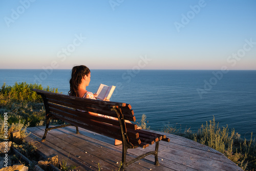 young woman sitting on a bench and reading book outdoors with summer sea background