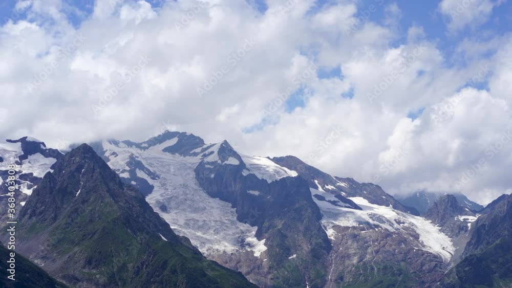 Snowy mountain against cloudy sky. From below white clouds floating on blue sky over mountain ridge covered with snow on sunny day in nature Snowy mountain against cloudy sky. From below white clouds floating on blue sky over mountain ridge covered with snow on sunny day in nature