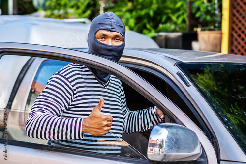 The male thief wears a black and white striped shirt and wears a black mask, smiling happily after stealing the car and getting a briefcase with money he wants.