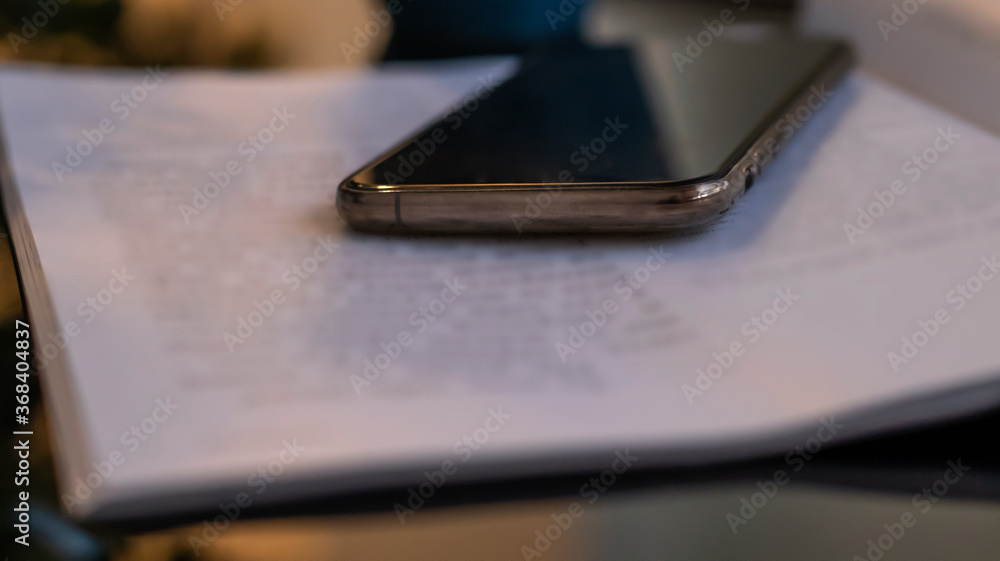 Open book and phone on a glass black table. Blurred boke background, close-up. Dark evening background with sunset highlights. Open book and phone on a glass black table. Blurred boke background, close-up. Dark evening background with sunset highlights.