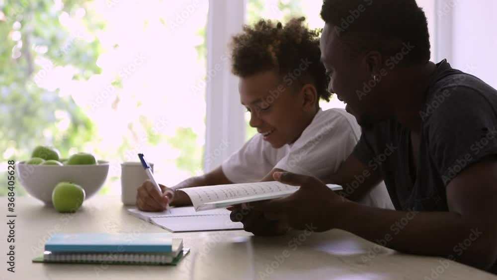 African American man and boy doing homework and talking at table in apartment interior spbi. Cheerful dad helping his son to learning, child writing in notebook and smiling, sitting at desk in bright African American man and boy doing homework and talking at table in apartment interior spbi. Cheerful dad helping his son to learning, child writing in notebook and smiling, sitting at desk in bright