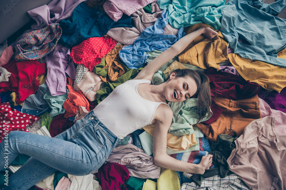 High angle above view photo of cheerful funny lady spring cleaning household lying among many clothes heap stack floor shopper surrounded sales stuff do not care mess spread hands indoors