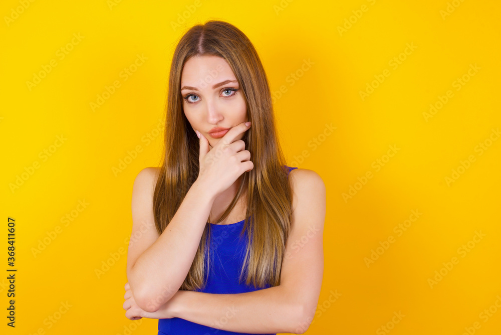 Portrait of thoughtful smiling girl keeps hand under chin, looks directly at camera, listens something with interest, dressed casually, poses against gray wall. Youth concept. Portrait of thoughtful smiling girl keeps hand under chin, looks directly at camera, listens something with interest, dressed casually, poses against gray wall. Youth concept.