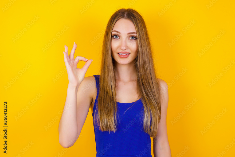 Glad attractive woman shows ok sign with hand as expresses approval, has cheerful expression, being optimistic. Standing against white wall. Glad attractive woman shows ok sign with hand as expresses approval, has cheerful expression, being optimistic. Standing against white wall.