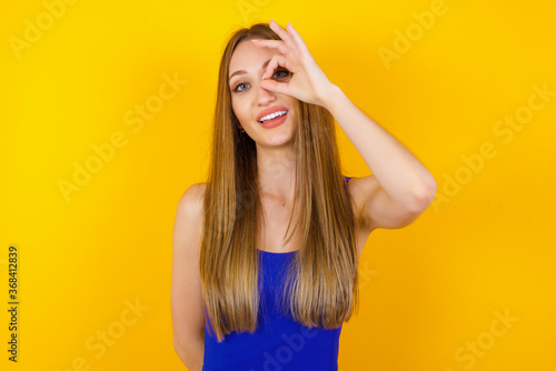 Beautiful young blonde woman with happy face smiling doing ok sign with hand on eye looking through finger. Wearing casual clothes and standing.